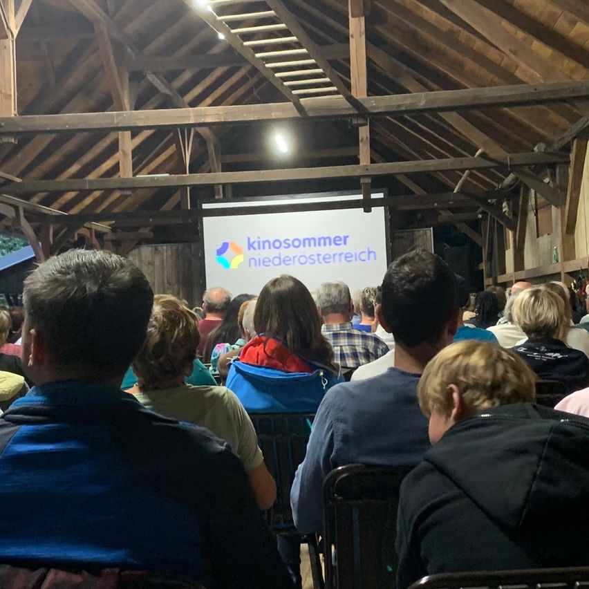 In einer Scheune zeigt eine Projektionsleinwand das Kinosommer Niederosterreich-Logo. Ein Publikum sitzt in Stühlen und schaut sich die Präsentation an, manche mit dem Rücken gedreht.
