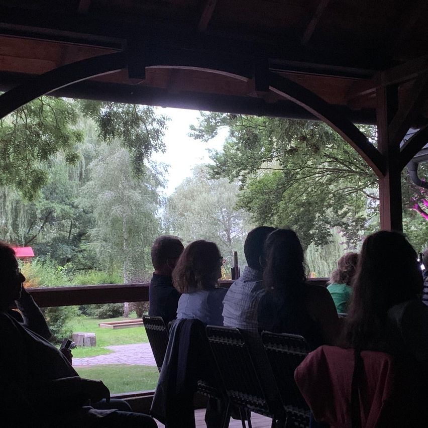 Eine Gruppe von Menschen sitzt in einem Pavillon und blickt auf eine üppige grüne Landschaft mit verschiedenen Bäumen. Die Umgebung erscheint ruhig und naturverbunden.