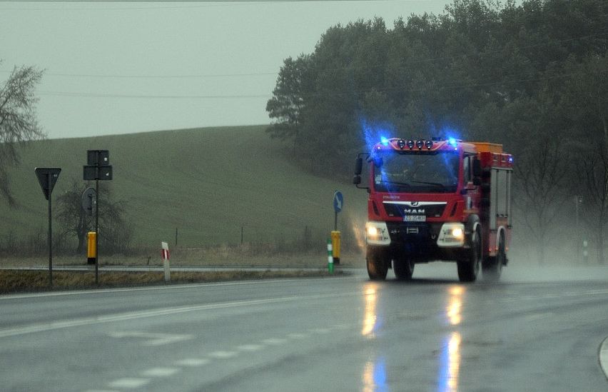 Ein roter Feuerwehrwagen mit blauen Lichtern fährt auf einer nassen Straße. Im Hintergrund sind Bäume und ein Hügel zu sehen.