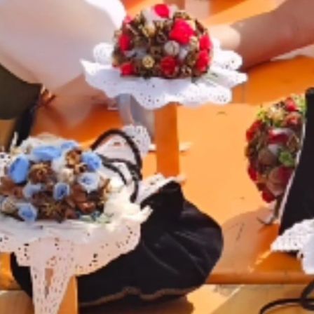 Ein orangefarbener Tisch zeigt verschiedene Gegenstände, darunter einen Blumenstrauß, eine schwarze Tasche und zwei weiße Tassen. Eine Hand mit einem Ring hält den Strauß.