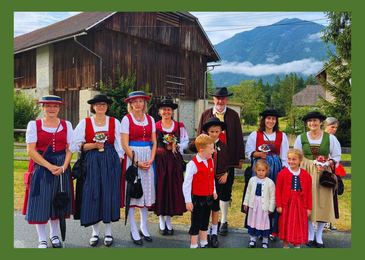 Eine Gruppe von Menschen in traditionellen Kostümen steht vor einer Holzscheune mit Bergen im Hintergrund. Einige tragen Blumen und halten Regenschirme.