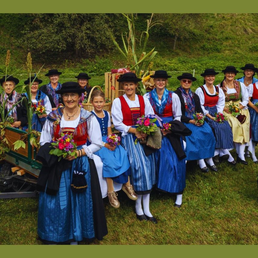 Eine Gruppe von Frauen und Mädchen in traditioneller Kleidung, die in einer Reihe auf einem Grasfeld stehen. Einige halten Blumen und Körbe. Ein Wagen steht hinter ihnen.