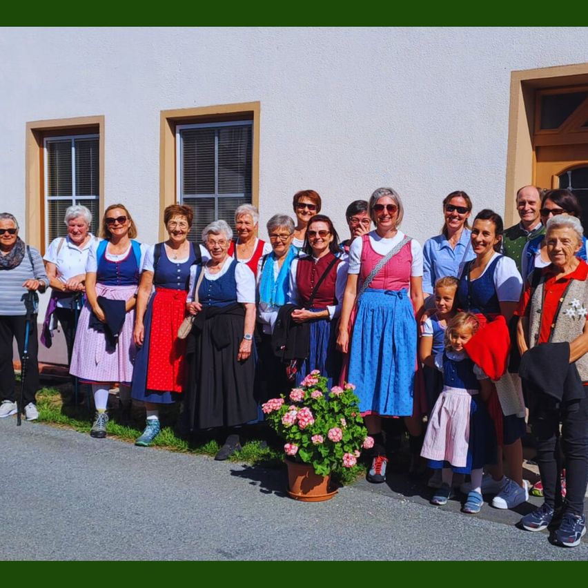 Eine Gruppe älterer Erwachsener, einige in traditioneller Kleidung, posiert für ein Foto vor einem Gebäude mit zwei Fenstern.