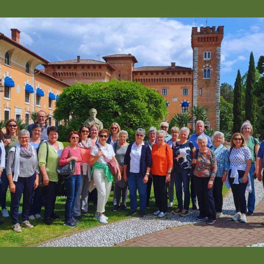 Eine Gruppe von Menschen steht vor einem Gebäude mit Turm, Büschen und einer Statue. Sie tragen Sonnenbrillen und verschiedene Kleidungsstücke.
