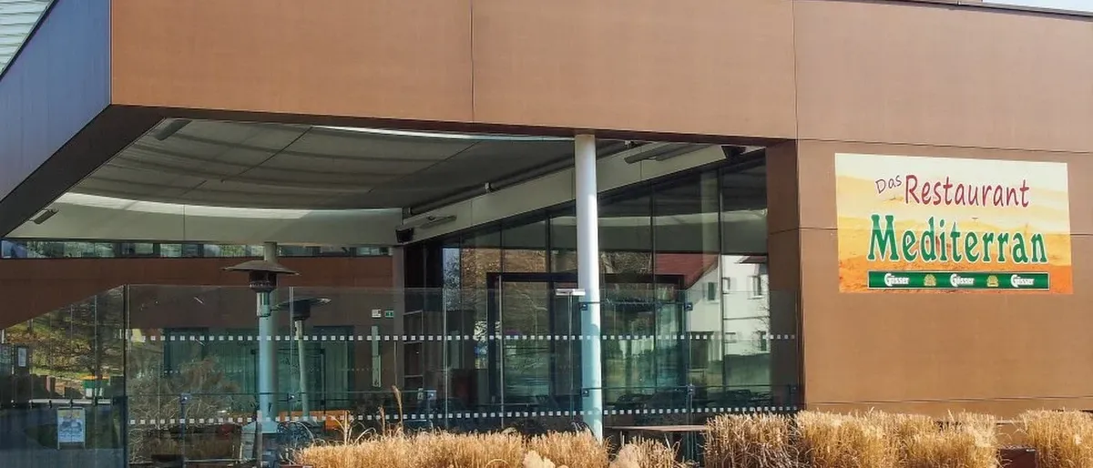 Der Eingang eines Gebäudes mit einer großen Glaswand, einem braunen Vordach und einem Schild mit dem Buchstaben M. Es gibt auch Glastüren und Fenster. Vor dem Gebäude stehen Pflanzen.