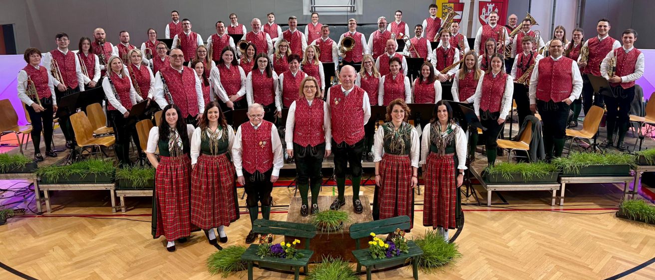 Eine Gruppe von Musikern in traditioneller Kleidung, die in einer Turnhalle für ein Foto posiert, mit einem Banner, das 'Stadtkapelle Freisach seit 1899' steht.