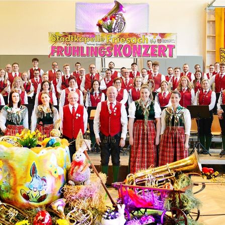 Ein Chor in traditionellen roten Westen und grünen Kleidern posiert mit Blechblasinstrumenten für ein Foto auf der Bühne. Ein Schild oben im Bild lautet 'Stadtkapelle Friesach Fruhlingskonzert'.