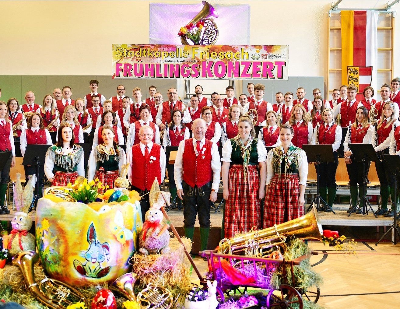 Ein Chor in traditionellen roten Westen und grünen Kleidern posiert mit Blechblasinstrumenten für ein Foto auf der Bühne. Ein Schild oben im Bild lautet 'Stadtkapelle Friesach Fruhlingskonzert'.