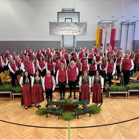 Eine Gruppe von Musikern in traditioneller Kleidung, einschließlich roter Westen und Röcke, posiert für ein Foto in einer Turnhalle mit einem Banner, das 'Stadtkapelle seit 1809 Freisach' lautet.