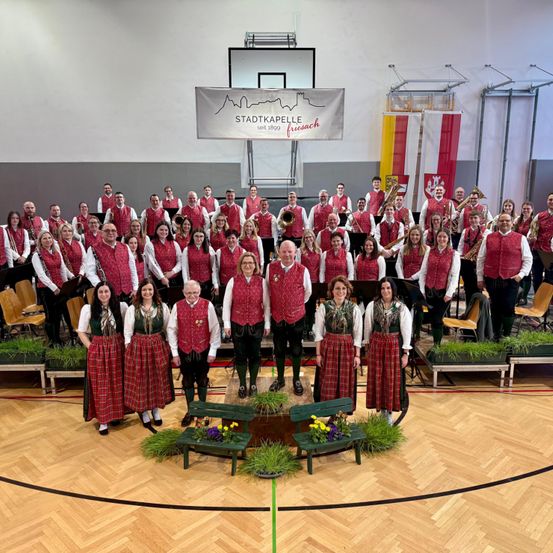 Eine Gruppe von Musikern in traditioneller Kleidung, einschließlich roter Westen und Röcke, posiert für ein Foto in einer Turnhalle mit einem Banner, das 'Stadtkapelle seit 1809 Freisach' lautet.
