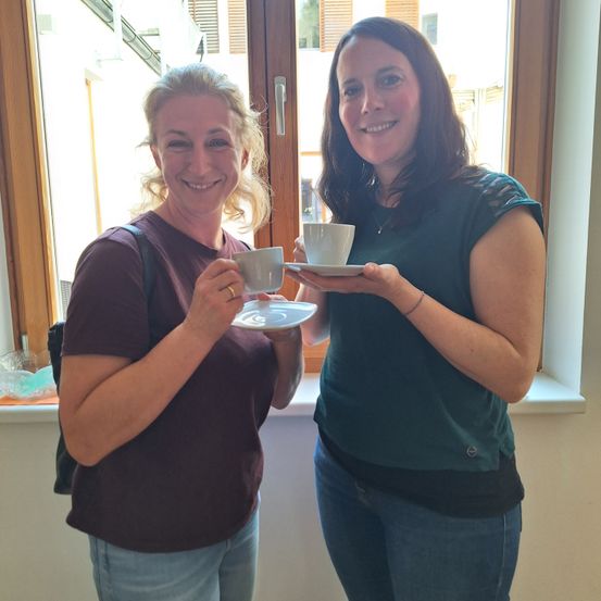 Zwei Frauen lächeln und halten Tassen mit Kaffee. Eine trägt ein braunes Shirt, die andere ein grünes Top. Sie stehen neben einem Fenster.