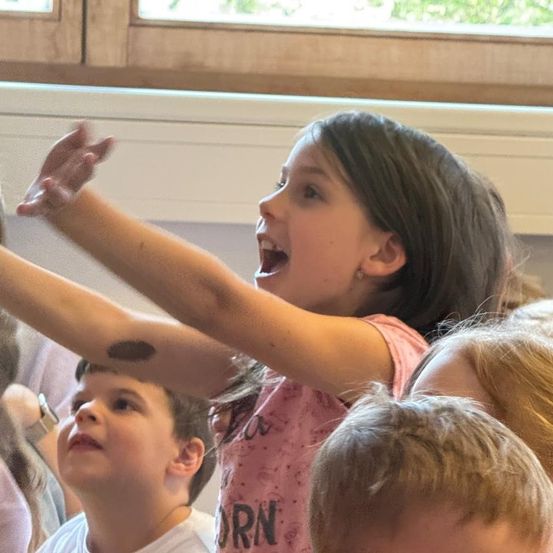 Ein Mädchen mit einem Tattoo am Arm hebt die Hände und singt. Andere Kinder beobachten sie.