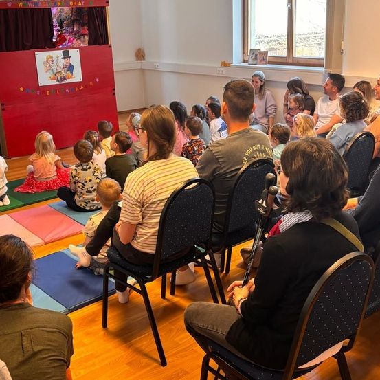 Eine Gruppe von Menschen schaut sich eine Puppentheateraufführung in einem Raum an. Das Publikum besteht aus Erwachsenen und Kindern, die auf Stühlen und Matten sitzen.