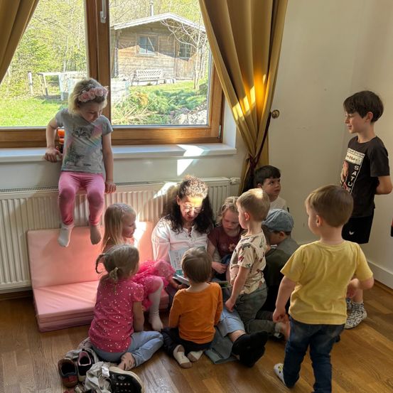 Eine Frau liest ein Buch vor einer Gruppe junger Kinder in einem Raum mit einem großen Fenster, das ein Haus überblickt.