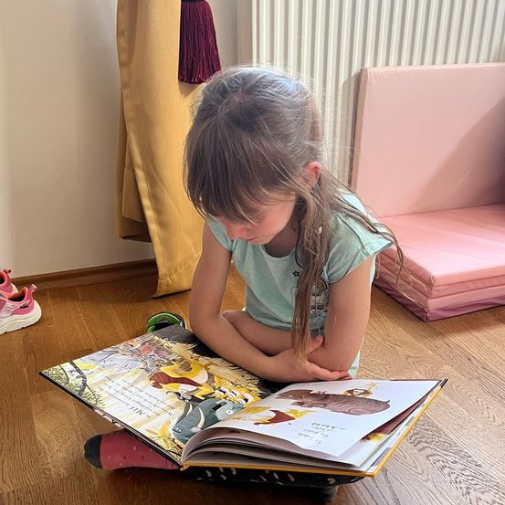 Ein junges Mädchen sitzt auf dem Boden, vertieft in ein Buch, mit verschränkten Beinen. Ein rosa gepolsterter Sitz ist hinter ihr. Ihre Turnschuhe liegen in der Nähe auf dem Boden.