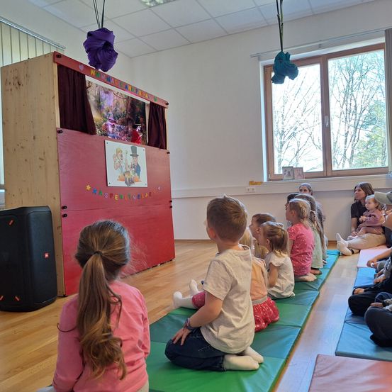 Eine Gruppe von Kindern sitzt auf grünen Matten und schaut sich eine Puppenshow auf einer roten Bühne mit bunten Dekorationen an. Erwachsene und Kinder sitzen im Raum mit Holzböden und weißen Wänden.