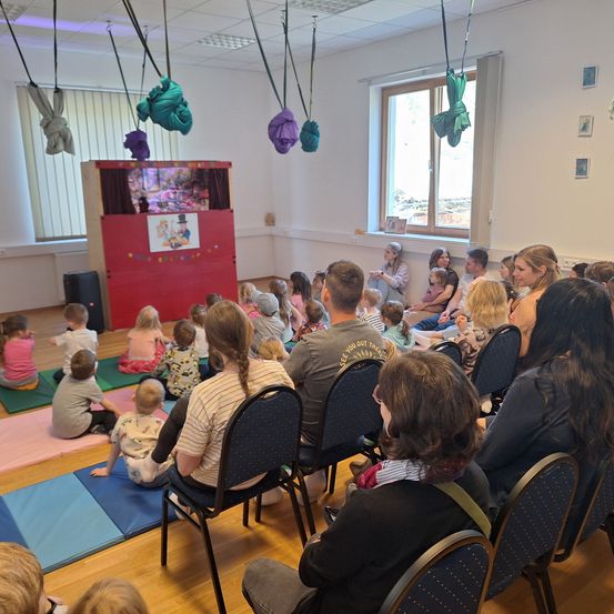 Eine Gruppe von Kindern und Eltern sitzt auf dem Boden und schaut sich eine Puppentheateraufführung auf einer kleinen Bühne an. Einige Spielzeuge hängen von der Decke.