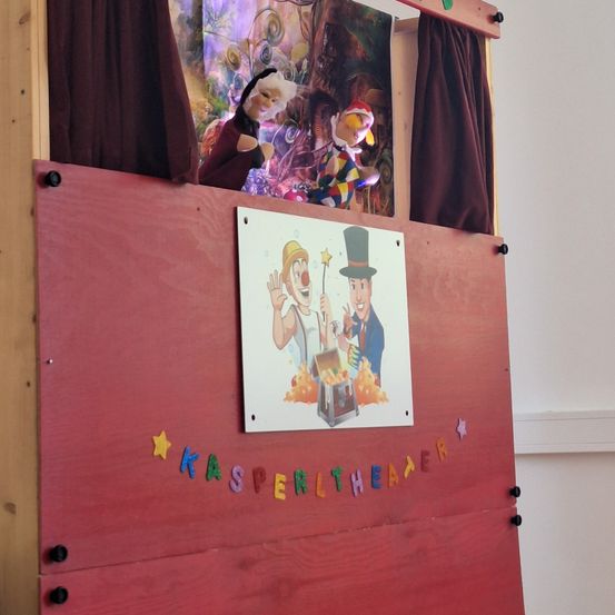 Ein rotes Puppentheater mit zwei Puppen hinter einem Vorhang. Ein Schild mit einem Clown-Bild und den Worten Kasperltheater auf der Vorderseite.