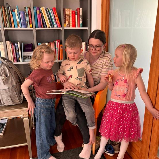 Eine Frau mit Brille sitzt auf einem Stuhl mit zwei Kindern. Eines liest ein Buch, das andere schaut zu. Sie sind von Büchern an Regalen umgeben.