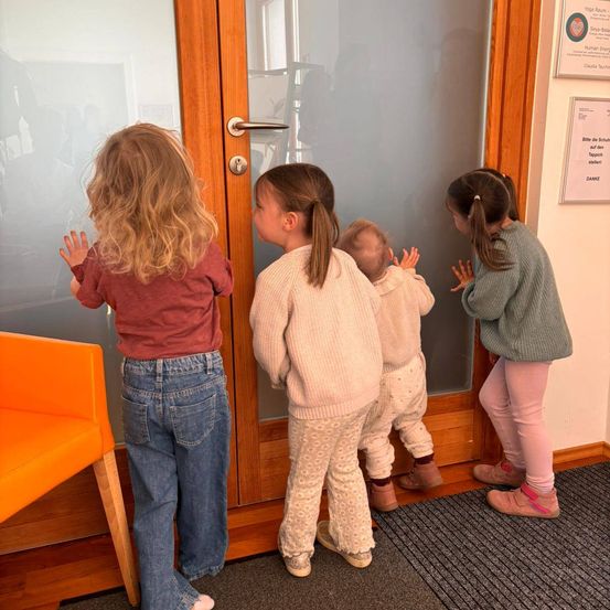 Vier Kinder, zwei Kleinkinder und zwei junge Mädchen, schauen aus einer Glastür. Sie scheinen auf jemanden zu warten. Das Mädchen auf der linken Seite hat ihre Hände auf die Glastür gelegt.