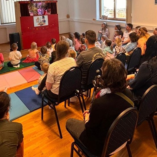Eine Gruppe von Kindern und Erwachsenen schaut sich eine Puppenshow auf einer Bühne in einem Raum mit Holzböden und Fenstern an.