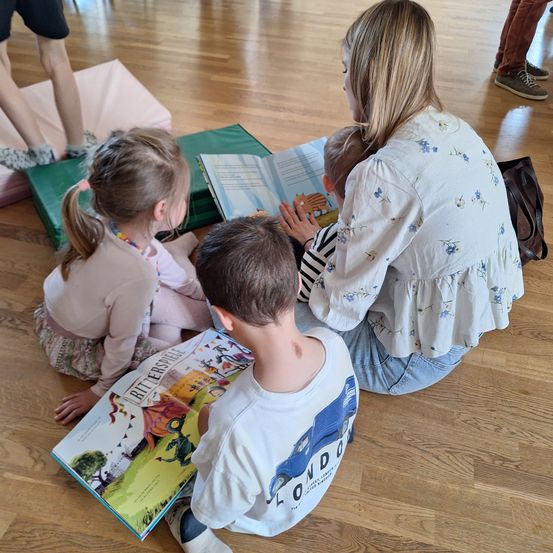 Eine Frau liest drei Kindern auf dem Boden sitzend ein Buch vor. Ein Kind hält ein Baby. Kissen und eine Handtasche sind in der Nähe.
