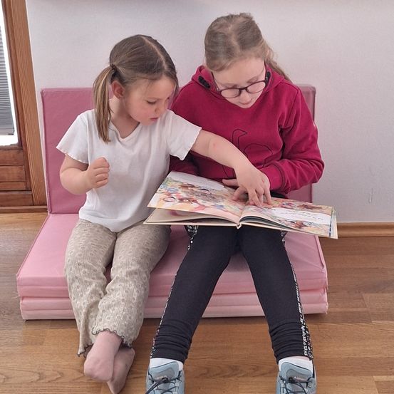 Zwei Mädchen sitzen auf einer rosa Bank. Eines liest ein Buch, während das andere zuhört. Der Raum hat Holzböden und eine weiße Wand.