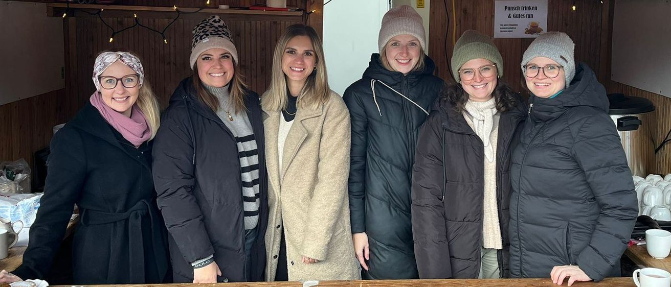 Eine Gruppe von Frauen steht hinter einem hölzernen Tresen in einer gemütlichen, hölzernen Hütte und lächelt für die Kamera. Über ihnen befinden sich Schilder, darunter eines für Kaffee, eines für Getränke und eines für Spaß.