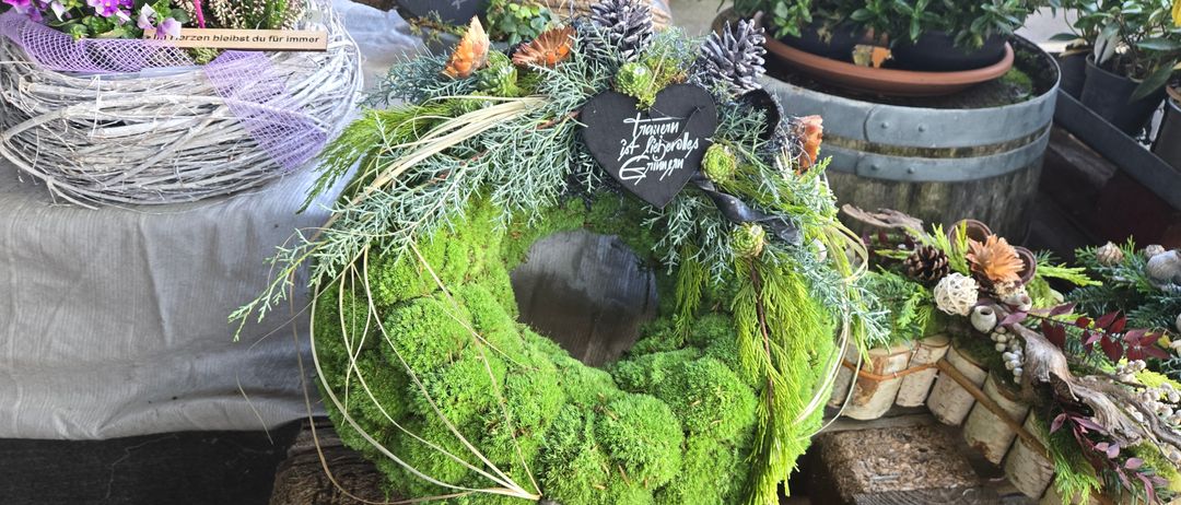 Ein Kranz aus Moos und Grünpflanzen steht auf einer Holzoberfläche. Er hat Tannenzapfen, Blätter und ein herzförmiges Schild mit den Worten '23. Liberelles Gringam'. Dahinter stehen Topfpflanzen und ein Korb.
