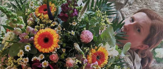 Eine Frau hält einen großen Blumenstrauß, darunter Rosen, Gänseblümchen und Sonnenblumen, mit grünen Blättern und einer Steinmauer im Hintergrund.