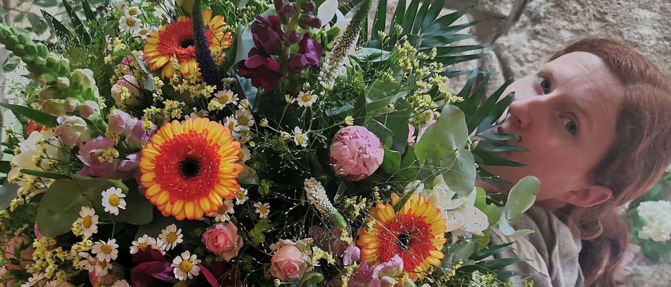 Eine Frau hält einen großen Blumenstrauß, darunter Rosen, Gänseblümchen und Sonnenblumen, mit grünen Blättern und einer Steinmauer im Hintergrund.