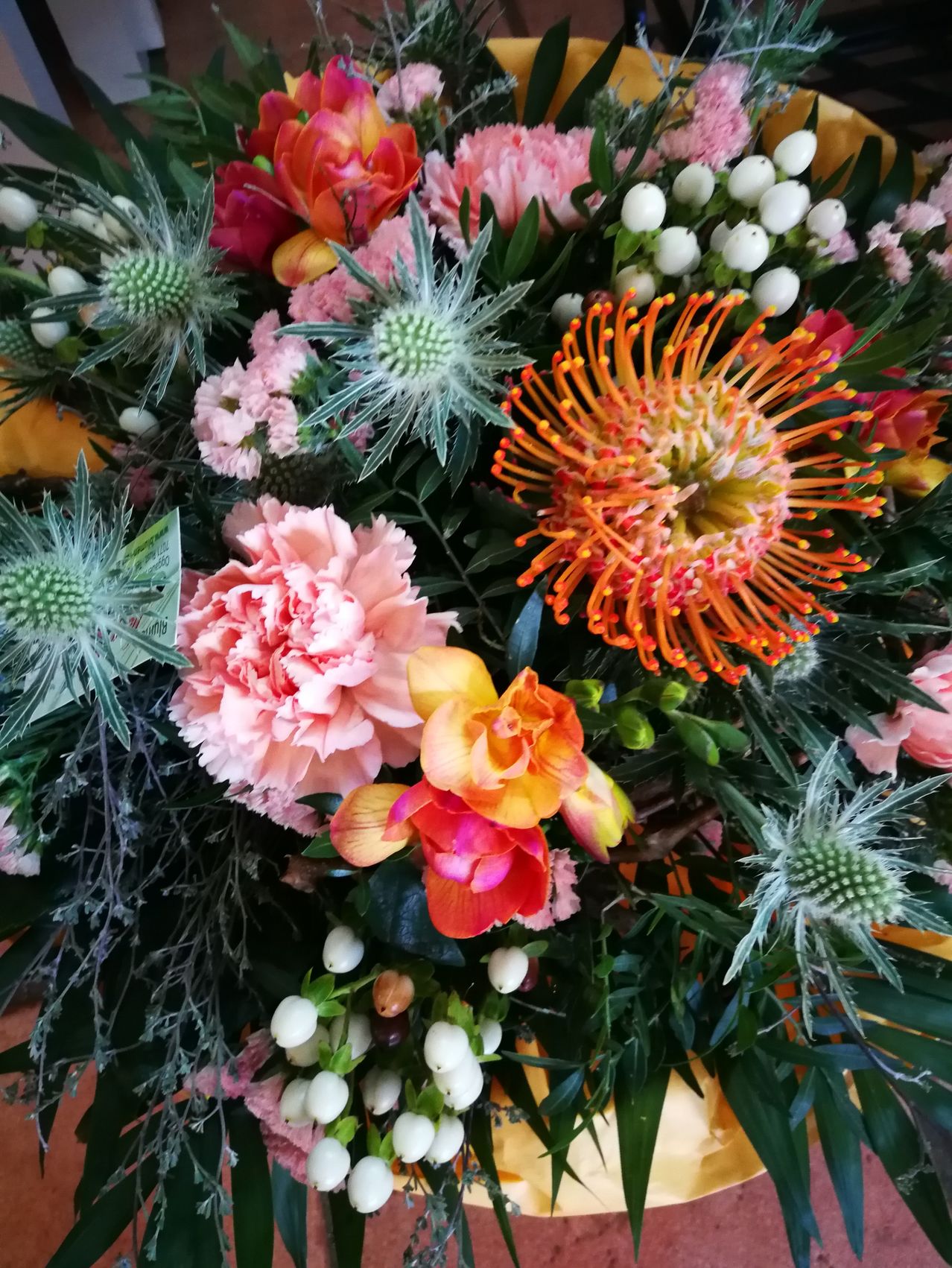Eine lebhafte Blumen-Arrangement mit rosa und orangefarbenen Blumen, spitzen grünen Blättern und kleinen weißen Knospen. Das Arrangement ist eine Mischung aus verschiedenen Blumenarten.