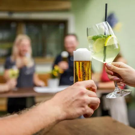 Zwei Hände stoßen mit einem Bier und einem Mojito an, mit verschwommenen Personen im Hintergrund, in einem Restaurant.