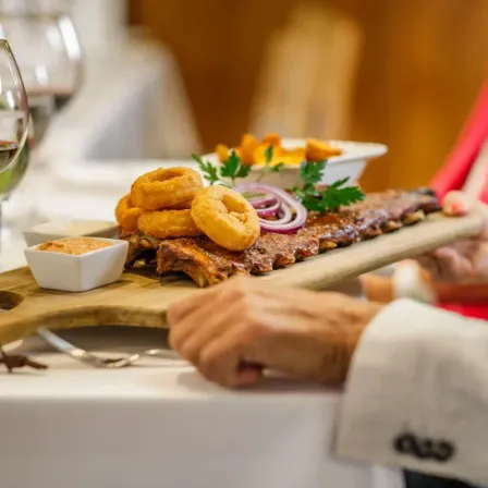 Ein Mann hält ein Schneidebrett mit Rippen und Zwiebelringen. Eine Schüssel mit Sauce und Weingläser stehen auf dem Tisch.