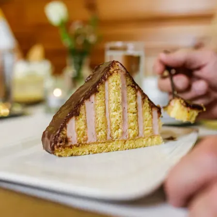 Eine Person schneidet in einen mit Schokolade überzogenen Kuchen mit Schinkenscheiben im Inneren. Der Kuchen befindet sich auf einem weißen Teller mit unscharfem Hintergrund.