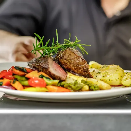Ein weißer Teller mit einem Steak, Gemüse und Kartoffeln, mit frischem Rosmarin garniert, serviert von einer Person in Schwarz.