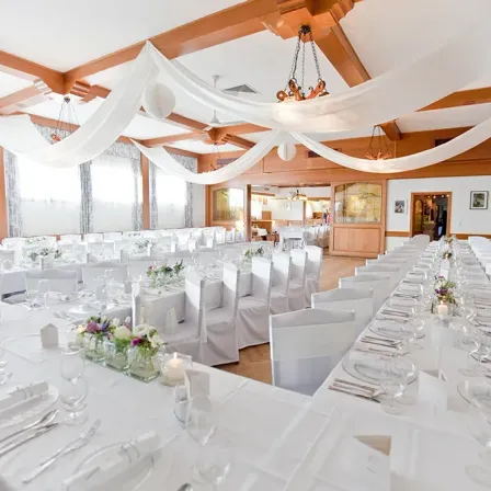 Ein Bankettsaal ist für eine formelle Veranstaltung vorbereitet. Tische sind mit weißen Tüchern, Blumen und Besteck geschmückt. Weiße Stühle sind ordentlich aufgestellt. Eine Holzdecke mit weißem Stoff und Kronleuchtern verleiht Eleganz.