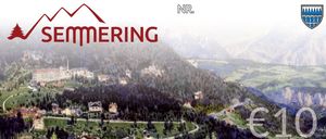 Ein malerisches Luftbild einer Bergregion mit dem Logo von NR. MERRING, das einen Tal, eine Straße und einige Gebäude mit Bäumen und Bergen in der Ferne zeigt.