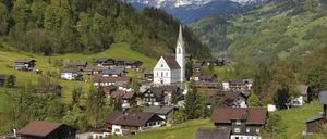 Ein malerisches Dorf in den österreichischen Alpen mit einer weißen Kirche, umgeben von grünen Feldern, Häusern und Blick auf schneebedeckte Berge.