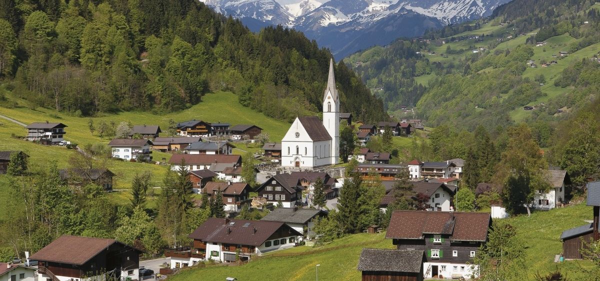 Ein malerisches Dorf in den österreichischen Alpen mit einer weißen Kirche, umgeben von grünen Feldern, Häusern und Blick auf schneebedeckte Berge.