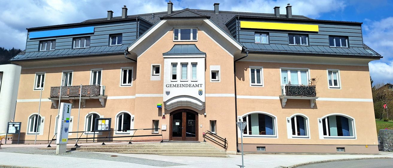 Ein mehrstöckiges beiges Gebäude mit grauem Dach und gelbem Schild, das Gemeindeamt heißt. Mehrere Fenster und eine Tür sind sichtbar.