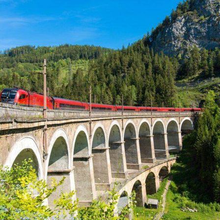 Ein roter Zug fährt über eine steinerne Bogenbrücke in einem bewaldeten Tal an einem sonnigen Tag.