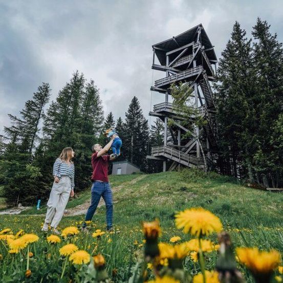 Eine dreiköpfige Familie spielt an einem sonnigen Tag auf einem Feld mit gelben Blumen und einem hölzernen Turm im Hintergrund.