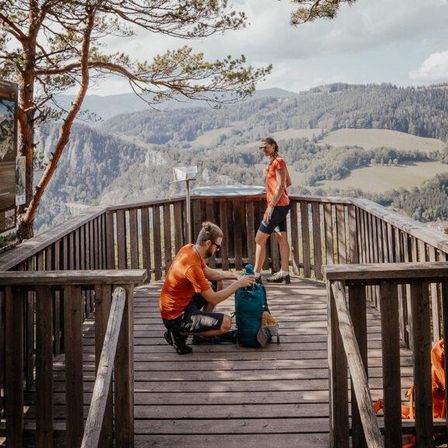 Ein Mann kniet auf einer Holzplattform und packt seinen Rucksack. Eine weitere Person steht in der Nähe und blickt auf die malerische Berg- und Tallandschaft hinunter.