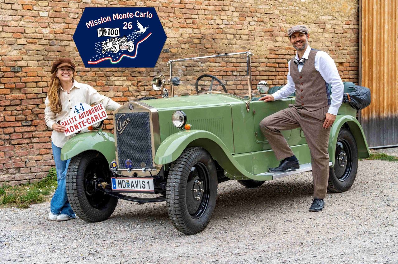 Zwei Männer in Vintage-Kleidung stehen neben einem grünen Oldtimer mit einem Schild, das 'Mission Monte-Carlo 26' steht. Das Auto hat ein Nummernschild, das 'MD AVIS 1' liest.