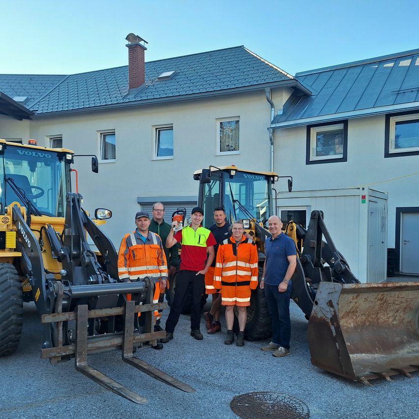 A group of people in high-visibility vests stand in front of a building with three large construction vehicles.