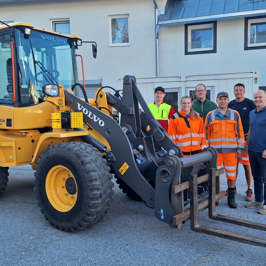 A group of workers in safety gear stand next to a yellow Volvo wheel loader in front of a building.