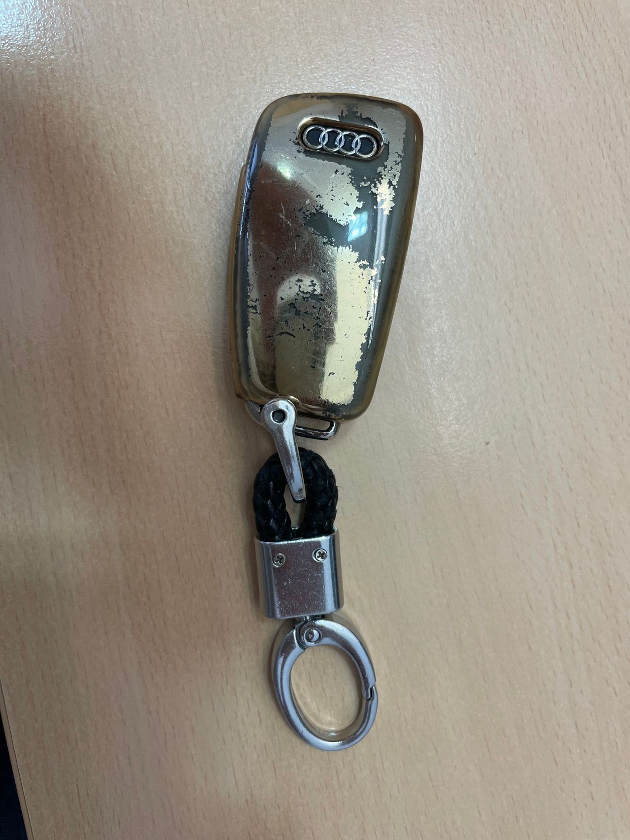 A metallic Audi car key fob with a black leather strap is placed on a wooden surface. The key fob has a silver ring and a black strap.
