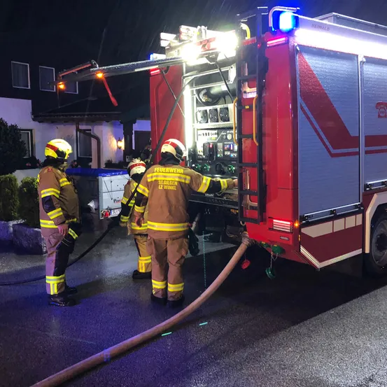 Drei Feuerwehrleute in voller Ausrüstung bedienen einen Feuerwehrwagen bei Nacht. Ein Feuerwehrmann klettert auf die Leiter des Fahrzeugs, während die anderen beiden daneben stehen.
