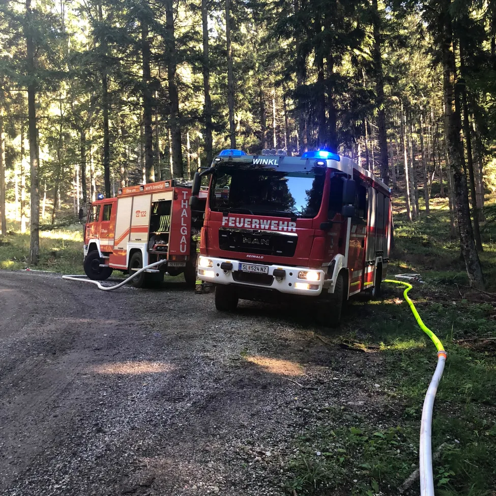 Zwei Feuerwehrfahrzeuge sind auf einem Schotterweg im Wald geparkt. Die Fahrzeuge haben ihre Lichter eingeschaltet und Schläuche angeschlossen.